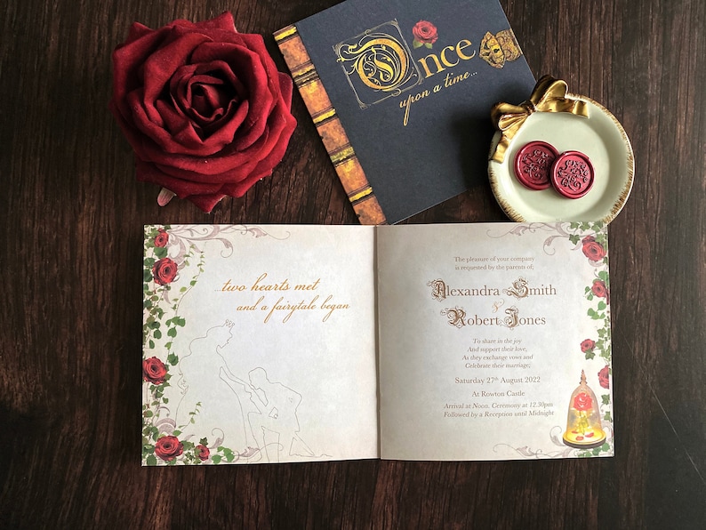May include: A wedding invitation suite with a fairytale theme. The open invitation features a silhouette of a couple and the text "two hearts met and a fairytale began." A red rose, a book, and a plate with wax seals complete the scene.