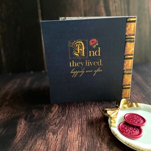 May include: A navy blue book with gold lettering that reads "And they lived, happily ever after." The book has a decorative spine and a red rose illustration. Two red wax seals are on a white and gold dish.