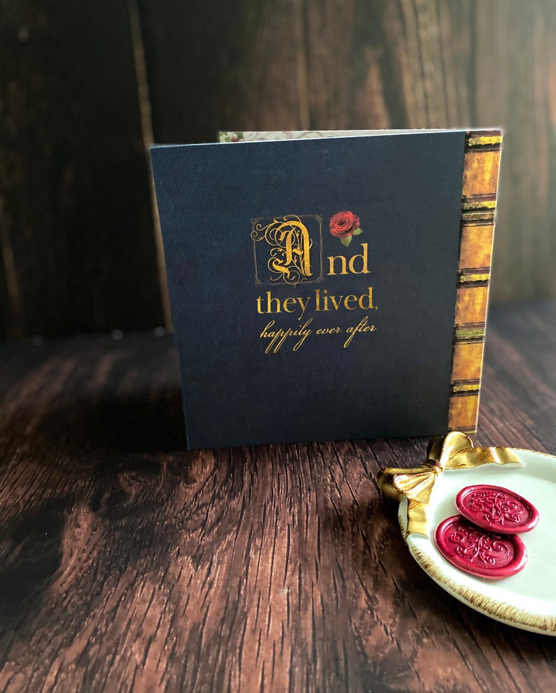 May include: A navy blue book with gold lettering that reads "And they lived, happily ever after." The book has a decorative spine and a red rose illustration. Two red wax seals are on a white and gold dish.