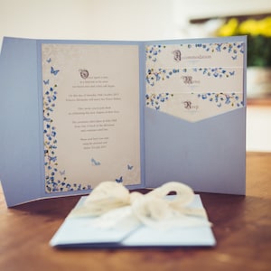 Può includere: Un invito di nozze blu chiaro con un design floreale e una tasca per carte aggiuntive. L'invito include i dettagli sulla cerimonia di nozze, il ricevimento e l'alloggio. Il testo sull'invito recita "Alloggio, Menu e RSVP."