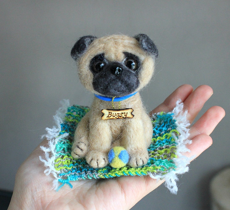 May include: A handmade felt pug dog figurine with a blue collar and a wooden name tag that reads "Bugsy." The pug is sitting on a small, colorful rug with a blue, green, and white pattern, and a small ball. The pug's eyes are dark and expressive.