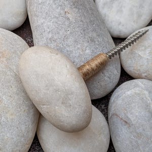 May include: A collection of smooth, rounded white and light gray river stones arranged together. One of the stones features a metal screw protruding from its center, with a small section of brown twine wrapped around the base of the metal screw.