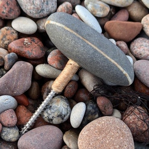 May include: A grey and white stone bottle stopper with a wooden screw. The stopper is lying on a bed of smooth, grey, brown, and red stones.