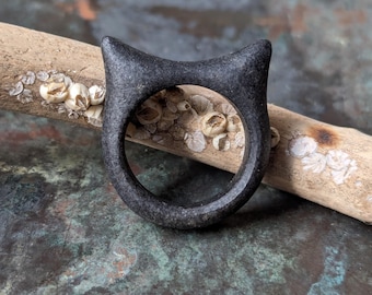 Puede incluir: Un anillo gris oscuro con diseño de orejas de gato. El anillo está hecho de un material sólido y tiene un acabado liso y mate. El anillo está sobre un trozo de madera flotante.