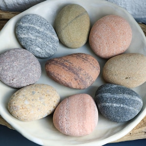 Può includere: Una ciotola di ceramica bianca piena di ciottoli lisci e colorati. I ciottoli sono di varie tonalità di grigio, marrone, rosa e beige.