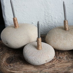 May include: Four smooth, light brown river rocks with a screw and twine wrapped around the top of each. The rocks are arranged on a piece of weathered wood.