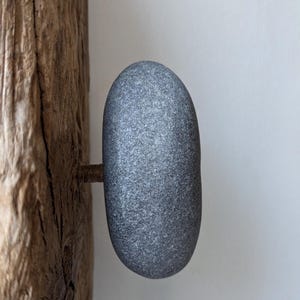 Könnte beinhalten: Ein grauer Stein-Schrankknauf mit einem Metallstiel, der an einem verwitterten Holzpfosten befestigt ist. Der Knauf ist oval geformt und hat eine strukturierte Oberfläche. Das Holz hat ein natürliches, gealtertes Aussehen.