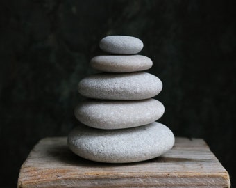 Könnte beinhalten: Ein Stapel aus fünf glatten, grauen Steinen, die auf einer Holzoberfläche übereinander balanciert sind. Die Steine sind in Pyramidenform angeordnet.