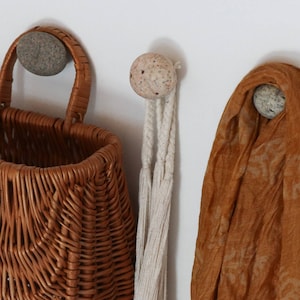 May include: A brown wicker basket with a handle hangs on a wall, next to a cream-colored macrame and a draped orange scarf. The items are suspended from stone-like wall hooks.