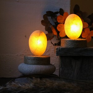 May include: Two glowing salt lamps, one larger than the other, are lit and placed on wooden and stone bases. The lamps are oval shaped and emit a warm, orange glow. The lamps are surrounded by dried foliage.