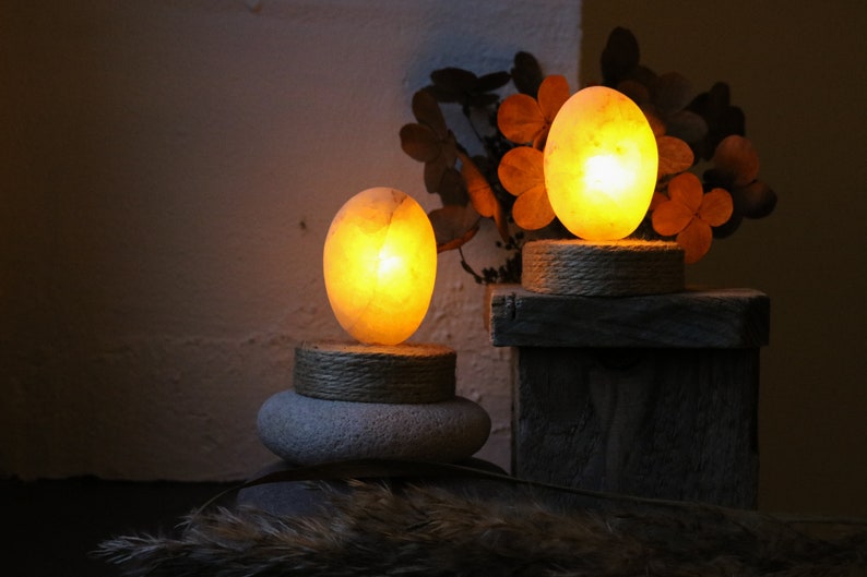 May include: Two glowing salt lamps, one larger than the other, are lit and placed on wooden and stone bases. The lamps are oval shaped and emit a warm, orange glow. The lamps are surrounded by dried foliage.