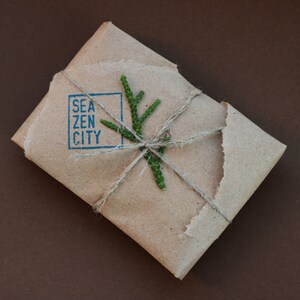 K&ouml;nnte beinhalten: Ein braunes Papierpaket, das mit Schnur gebunden und mit einem kleinen gr&uuml;nen Zweig verziert ist. Das Paket ist mit den Worten "Sea Zen City" in einem blauen Quadrat gestempelt.