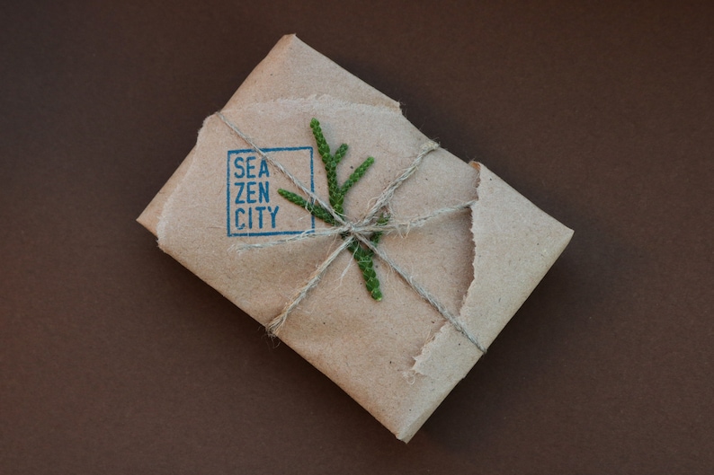 K&ouml;nnte beinhalten: Ein braunes Papierpaket, das mit Schnur gebunden und mit einem kleinen gr&uuml;nen Zweig verziert ist. Das Paket ist mit den Worten "Sea Zen City" in einem blauen Quadrat gestempelt.
