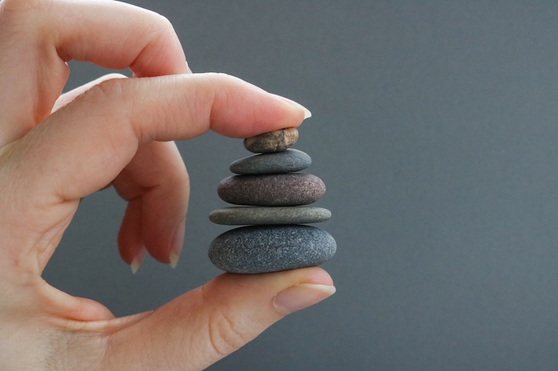 K&ouml;nnte beinhalten: Eine Hand balanciert vorsichtig einen Stapel aus f&uuml;nf glatten, grauen und braunen Steinen. Die Steine sind pyramidenf&ouml;rmig gestapelt, wobei der kleinste Stein oben liegt.