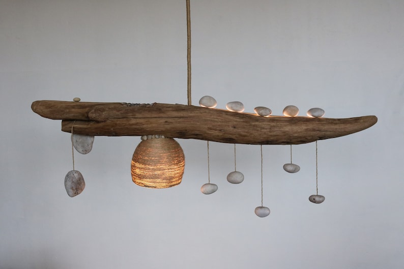 May include: A rustic wooden branch chandelier with a woven paper shade and multiple white stones hanging from it. The stones are suspended from the branch by strings.