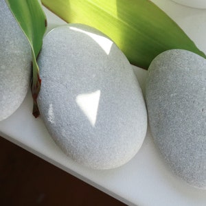 May include: Three smooth, gray stones arranged on a white surface. A green leaf partially covers the stones, casting a shadow on the middle stone.
