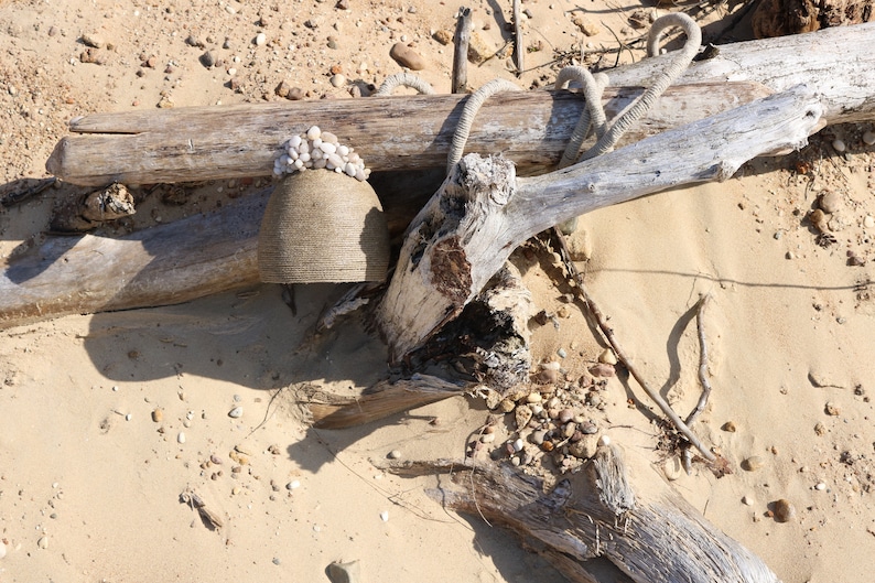 May include: A weathered, gray wooden log with a rope wrapped around it, resting on a sandy beach. A smaller, gray, ceramic pot with white shells glued to the top sits on the log.