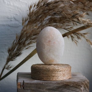 May include: A white egg-shaped stone sits on a round base made of natural twine. The base is on a piece of weathered wood. A dried grass plume is in the background.