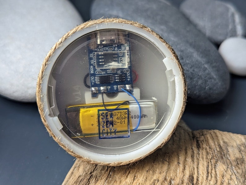 May include: A small, round, clear plastic container with a rope border. Inside the container is a small circuit board with wires and a yellow battery labeled "3.7V 450mAh 400mAh SEW ZEN CITY 42-01".