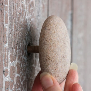 May include: A light brown, oval-shaped stone cabinet knob. It is attached to a wooden surface with a metal rod. The wood has a distressed appearance. The knob is likely made of natural stone.