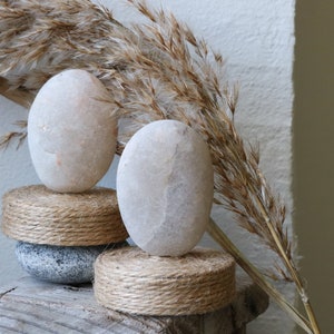 May include: Two smooth, white stones are stacked on top of two round, woven rope bases. The stones are on a weathered wooden surface with a dried, brown grass plant in the background.