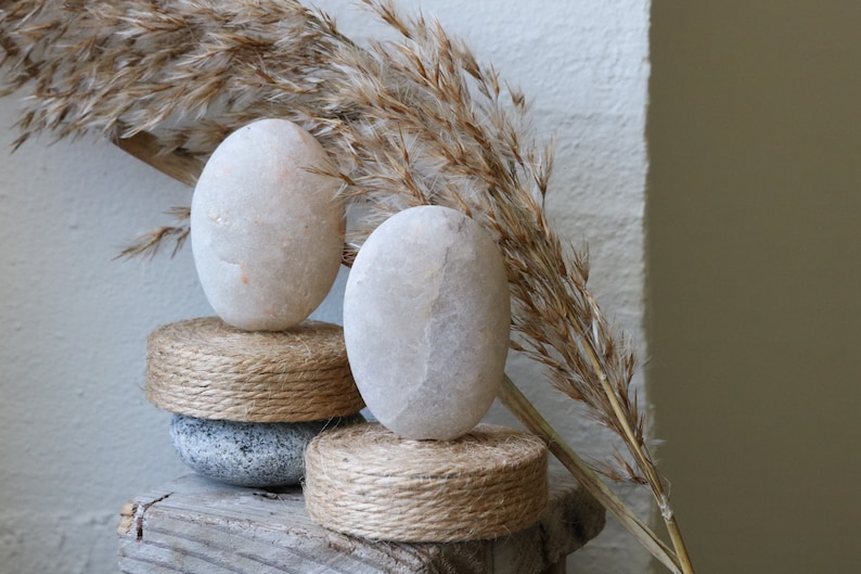 May include: Two smooth, white stones are stacked on top of two round, woven rope bases. The stones are on a weathered wooden surface with a dried, brown grass plant in the background.