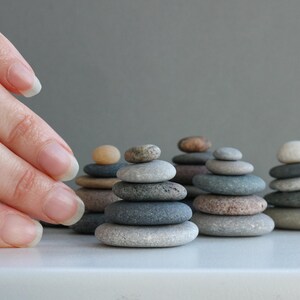 K&ouml;nnte beinhalten: Eine Hand greift nach einer Reihe kleiner Steinhaufen, die jeweils aus glatten, grauen, braunen und wei&szlig;en Steinen bestehen. Die Haufen sind in einer Reihe auf einer wei&szlig;en Oberfl&auml;che angeordnet.