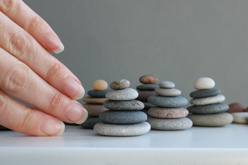 K&ouml;nnte beinhalten: Eine Hand greift nach einer Reihe kleiner Steinhaufen, die jeweils aus glatten, grauen, braunen und wei&szlig;en Steinen bestehen. Die Haufen sind in einer Reihe auf einer wei&szlig;en Oberfl&auml;che angeordnet.