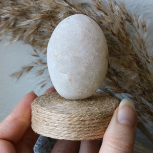 May include: A white, smooth, oval-shaped stone sits on a small, round, brown, woven base. The base is made of twine.