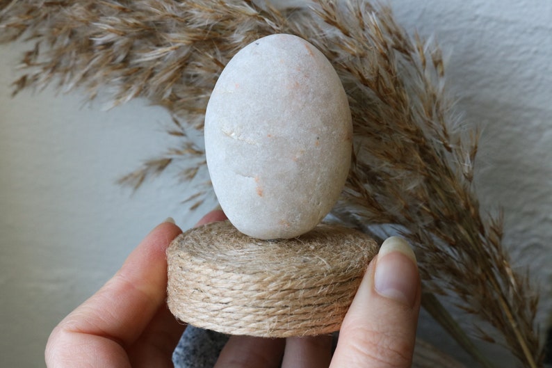 May include: A white, smooth, oval-shaped stone sits on a small, round, brown, woven base. The base is made of twine.