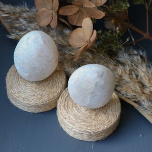 May include: Two white stone eggs on natural twine bases. The eggs are smooth and round, and the bases are made of natural twine. The eggs are sitting on a dark blue surface.