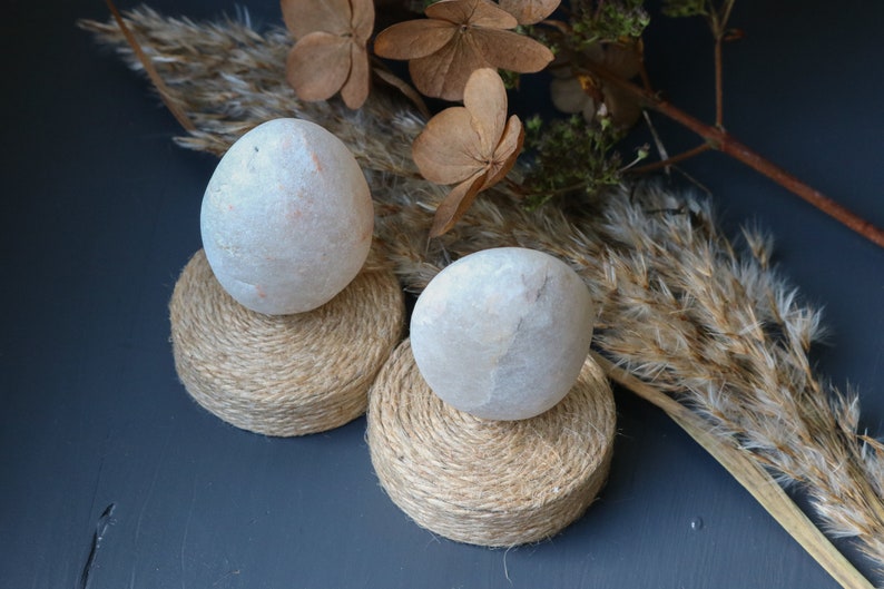 May include: Two white stone eggs on natural twine bases. The eggs are smooth and round, and the bases are made of natural twine. The eggs are sitting on a dark blue surface.