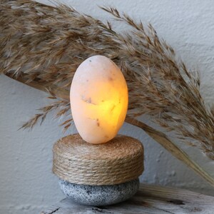 May include: A warm, glowing salt lamp in the shape of an egg, sitting on a natural woven base and a gray stone base. The lamp is illuminated with a soft, orange light.