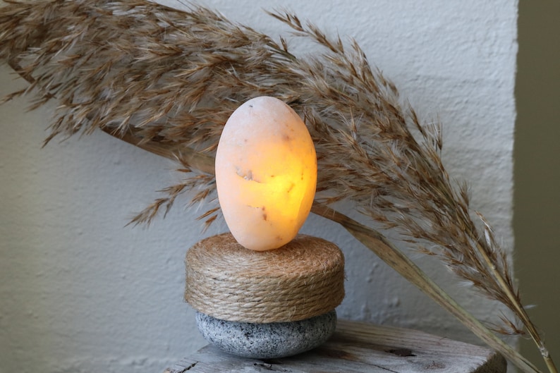 May include: A warm, glowing salt lamp in the shape of an egg, sitting on a natural woven base and a gray stone base. The lamp is illuminated with a soft, orange light.