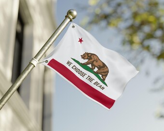 May include: A California state flag waving in the wind. The flag features a brown bear on a patch of green grass, a red star, and a red stripe at the bottom. The text "WE CHOOSE THE BEAR" is printed on the flag.