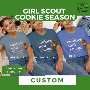 May include: Three women wearing different colored t-shirts with the text "Powered by cookies and chaos" and a cookie graphic. The text "TROOP 33459" is below the graphic. The t-shirts are in the colors stone blue, indigo blue, and iris. The text "STONE BLUE", "INDIGO BLUE", and "IRIS" is below each shirt. The text "ADD YOUR TROOP #" is below the shirts. The text "CUSTOM" is at the bottom of the image.