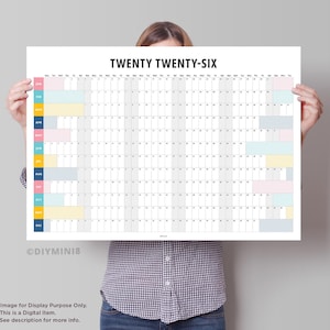 May include: A large white 2026 calendar with the text "TWENTY TWENTY-SIX" at the top. Months are listed vertically on the left with color-coded blocks. The calendar is held by a person.