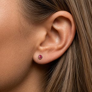 May include: A close-up of a person's ear with a small, round, ruby-colored stud earring. The earring is set in a silver or white gold bezel. The person's hair is a light brown color.