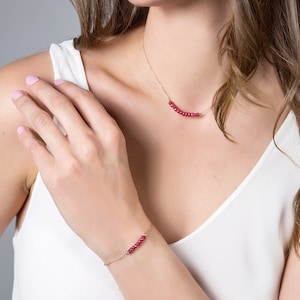 Può includere: Un set di collana e bracciale in oro con piccole pietre preziose rosse vibranti. La collana e il bracciale hanno un design delicato. I gioielli sono esposti su una persona che indossa un top bianco.