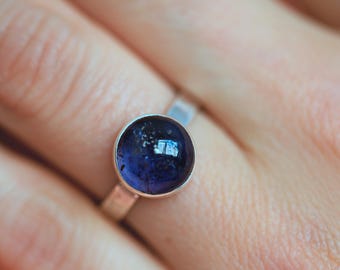 May include: A silver ring with a large, round, deep blue gemstone. The gemstone has a polished, reflective surface. The ring's band is a simple, polished silver. The ring is shown on a person's finger.