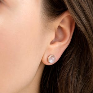 Op de afbeelding: Close-up van een oor met een ronde, lichtroze oorbel. De oorbel heeft een zilveren bezel setting. De persoon heeft bruin haar.