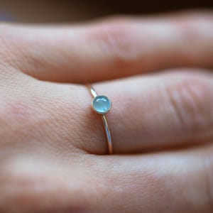 May include: A delicate gold ring featuring a small, round, light blue gemstone. The ring has a thin band and a simple, elegant design. The gemstone is set in a bezel setting, and the ring is shown on a finger.