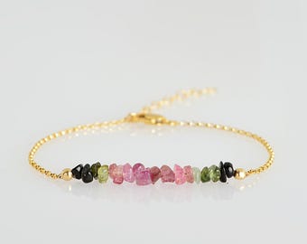 May include: A gold-coloured bracelet with a chain and a row of multi-coloured tourmaline gemstones. The gemstones are in shades of pink, green, and black. The bracelet has a delicate chain and a secure clasp.