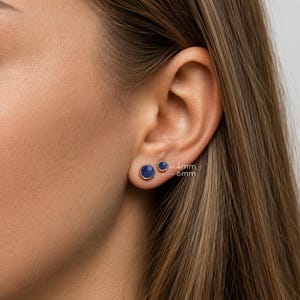 Puede incluir: Dos pendientes de botón con un diseño circular azul oscuro. El pendiente más pequeño tiene 4 mm de diámetro y el más grande 6 mm. Los pendientes están engastados en un metal color oro.