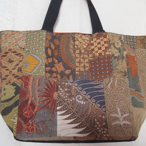 May include: A large tote bag with a patchwork design. The bag features a variety of patterned fabric squares in shades of brown, tan, blue, and red. It has black handles and a black base, suitable for carrying items.