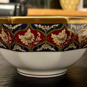 May include: A white and gold porcelain bowl with a detailed floral pattern in red, blue, and green. The rim is trimmed with gold. The bowl has a unique octagonal shape.
