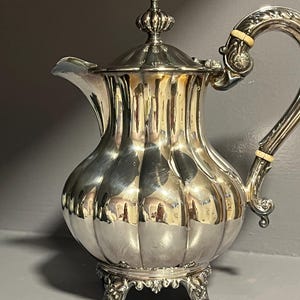 Op de afbeelding: Een gepolijste verzilverde theepot met een bolvormig, verticaal gegroefd lichaam, een gebogen handvat met lichtgekleurde isolatieringen, een decoratieve knop op het deksel en drie sierlijke, gekrulde voetjes onder de basis van de pot.