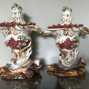 May include: Pair of ornate white porcelain vases with gold accents, burgundy details, and floral patterns. The vases have a unique shape with a flared top and a detailed base, showcasing a classic, elegant design.
