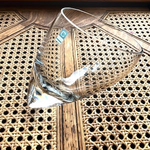 May include: A clear glass tumbler with a slanted, asymmetrical rim and a thick, pointed base. The glass features a small, square teal sticker with a white letter C on the upper side, showcasing a smooth, transparent surface and a distinct geometric shape.