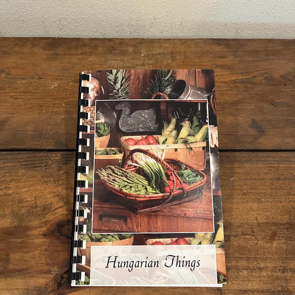 May include: A spiral bound cookbook with a white cover and the title "Hungarian Things" printed in black. The cover features a photograph of a basket filled with fresh vegetables, including asparagus, green onions, and red tomatoes.